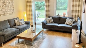 A living room with two sofas, a coffee table, and a TV. There are curtains, a lamp, and a view of the outdoors. - Family home near Darß—fireplace, patio, and plenty of space surrounded by nature (Pruchten)