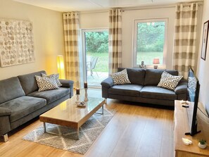 A living room with two sofas, a coffee table, and a TV. There are curtains, a lamp, and a view of the outdoors. - Family house near Darß - fireplace & terrace, quiet location, tastefully furnished. (Pruchten)