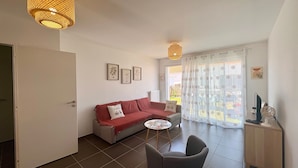 A living room with a red sofa, a small round table, a grey armchair, a wooden cabinet, and a large window with patterned curtains. - House between forest and beach - 4 guests (Saint-Jean-de-Monts)