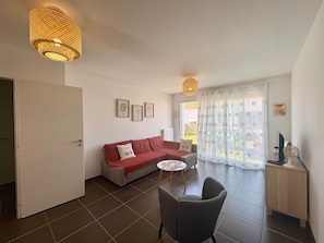 Ein Wohnzimmer mit rotem Sofa, einem kleinen runden Tisch, einem grauen Sessel, einer Holzkommode und einem großen Fenster mit gemusterten Vorhängen.