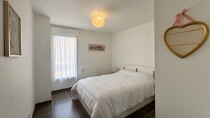 A bedroom with a bed, a window with curtains, a radiator, a wall-mounted picture frame, and a heart-shaped decoration. - House between forest and beach - 4 guests (Saint-Jean-de-Monts)