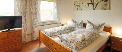 Une chambre avec un lit en bois, un téléviseur posé sur une commode, une fenêtre avec des rideaux et un mur décoré d’œuvres d’art florales.
