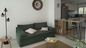 Ein Wohnzimmer mit einem grünen Sofa, einem kleinen Tisch, einer Stehlampe, einem dekorativen Spiegel und einer Küchenzeile mit Barhocker und Kühlschrank.