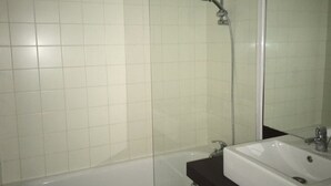 Un baño con paredes de azulejos blancos, una bañera, un lavamanos con espejo y un cabezal de ducha.