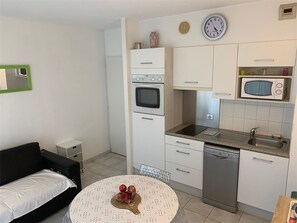 A compact kitchen with a microwave, oven, and dishwasher. There is a dining table with a fruit bowl and a black sofa. - Apartment with pool - 4 guests (Moliets-et-Maa)