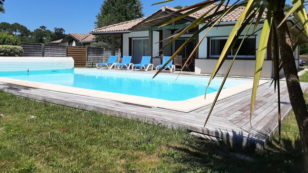 Une piscine avec des chaises longues, une terrasse en bois et une maison avec un toit en tuiles.