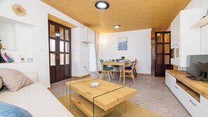 A compact living space with a wooden ceiling, a glass-top coffee table, a sofa, a dining area with chairs, and a TV on a wooden cabinet. - Family-friendly accommodation with terrace & garden (Tribalj)