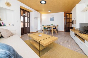Ein gemütlicher, kompakter Wohnraum mit Holzboden, einem Glastisch, einer Couch, einem Essbereich mit Stühlen und einem Fernseher auf einer Holzkommode.