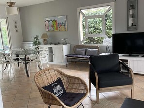 A modern living room with a dining area, a television, and a large window with a view of greenery. - House with heated pool - 8 guests (Moliets-et-Maa)
