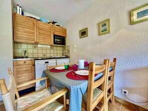 Eine kompakte Küche mit Holzausstattung, einer Mikrowelle und einem Geschirrspüler. Ein Esstisch mit Stühlen und ein kleines, an der Wand befestigtes Regal sind ebenfalls vorhanden.
