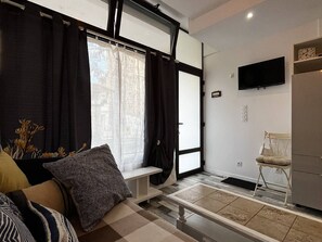 Ein modernes Wohnzimmer mit einer Couch, einer Bank, einem Sessel, einem Flachbildfernseher und einem großen Fenster mit Vorhängen.