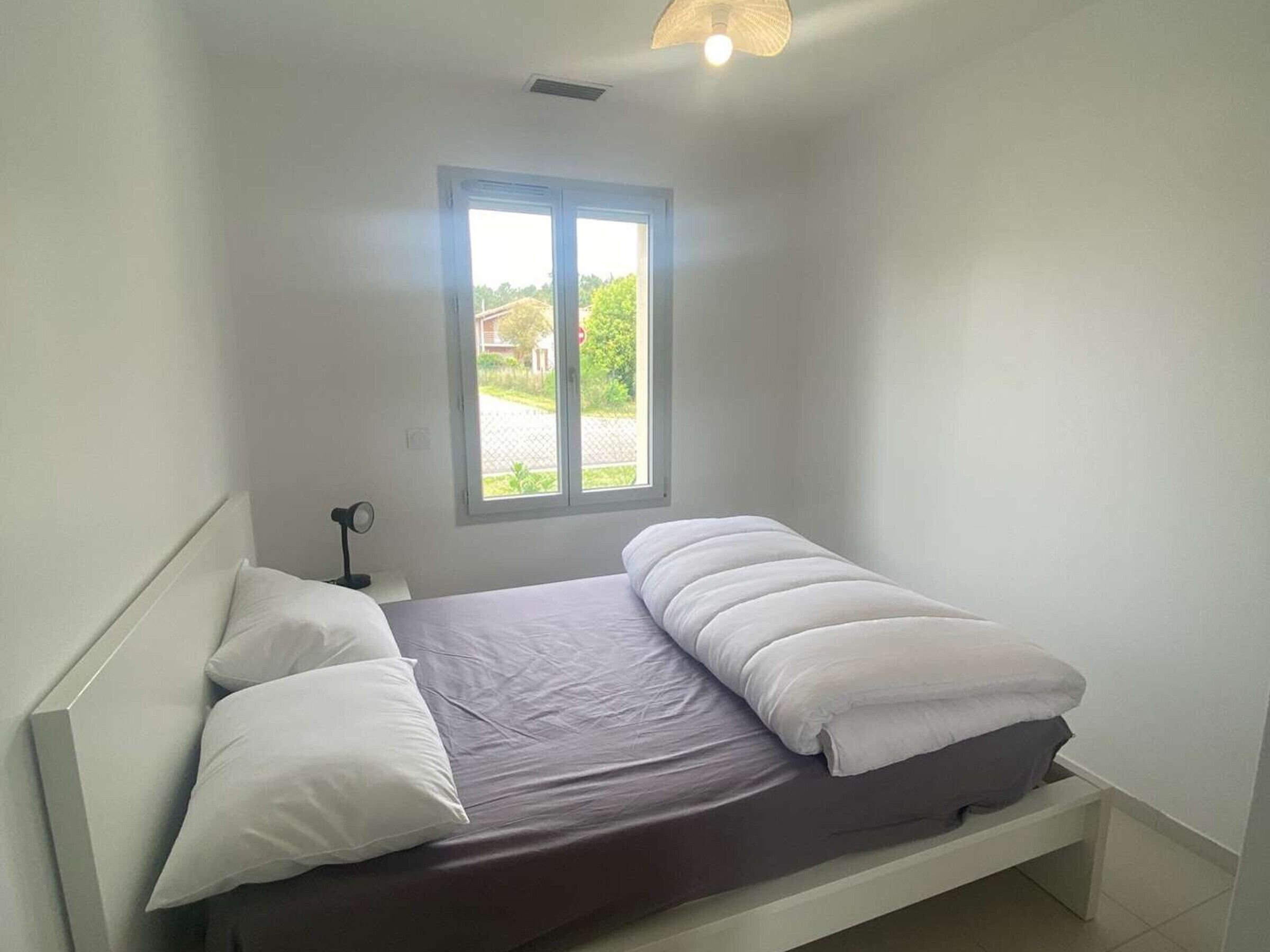 A single bed with white bedding, a nightstand with a lamp, and a window with a view of greenery.