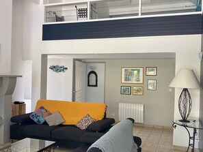 A living room with a black sofa, a yellow throw, a glass coffee table, and a black armchair. There is a glass table with a lamp, a radiator, and framed pictures on the wall. - Villa with private heated pool - 10 guests (Moliets-et-Maa)