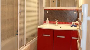 Casa de banho com cabine de chuveiro em vidro, toucador vermelho com bancada branca e espelho sobre a pia.