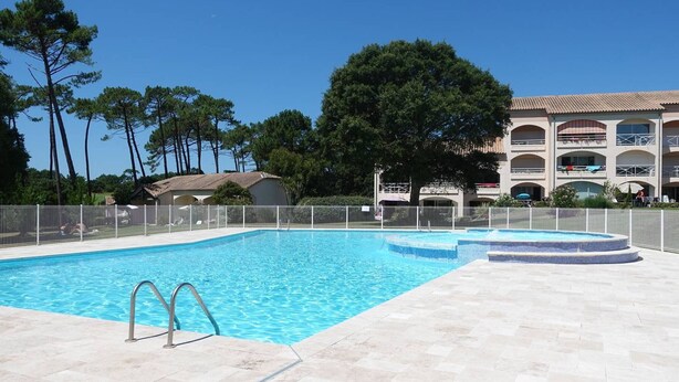 Piscina com deck revestido, cercada por uma grade, e um edifício com várias varandas ao fundo.