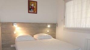 Quarto com cabeceira de madeira, duas luminárias de cabeceira, cama com roupa de cama branca, uma tomada e uma janela com cortina translúcida.