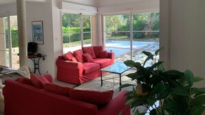 A spacious room with a red sofa, a glass coffee table, and large windows offering a view of a swimming pool and greenery. - Villa with pool - 10 guests (Moliets-et-Maa)
