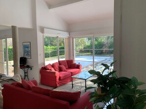 A spacious room with a red sofa, a glass coffee table, and large windows offering a view of a swimming pool and greenery. - Villa with pool - 10 guests (Moliets-et-Maa)