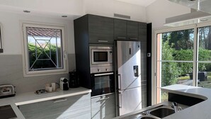 A modern kitchen with a built-in oven, microwave, and refrigerator, a window with a view of greenery, and a sink with a stainless steel faucet. - Villa with pool - 10 guests (Moliets-et-Maa)