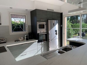 A modern kitchen with a built-in oven, microwave, and refrigerator, a window with a view of greenery, and a sink with a stainless steel faucet. - Villa with pool - 10 guests (Moliets-et-Maa)