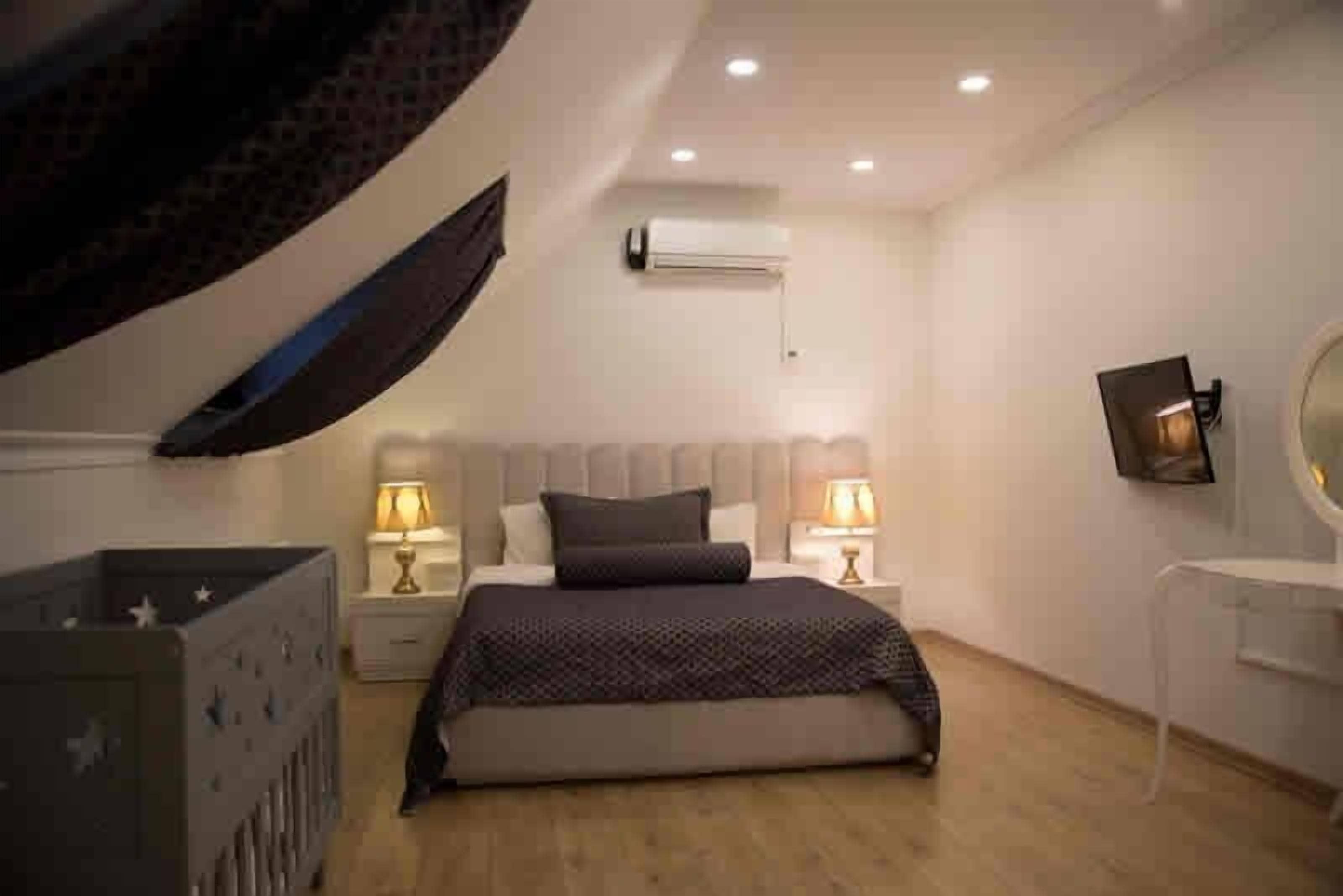 A bedroom with a bed, two bedside tables with lamps, a wall-mounted TV, and an air conditioning unit.