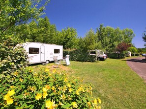 Un terrain de camping avec un camping-car blanc, une petite caravane blanche et une voiture garée sur une zone herbeuse, au milieu d’arbres et d’arbustes.