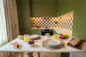 Ein Frühstückstisch mit Tellern, Brot, Obst und Saft auf einer weißen Tischplatte in einer Küche mit grünen Wänden und einer karierten Spritzschutzleiste.
