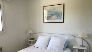 Un dormitorio con cama blanca, dos mesitas de noche con lámparas, un cuadro en la pared y una ventana a la izquierda.