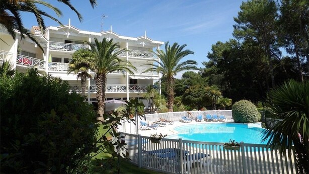Un hotel de varios pisos con piscina rodeada de palmeras y una valla blanca.