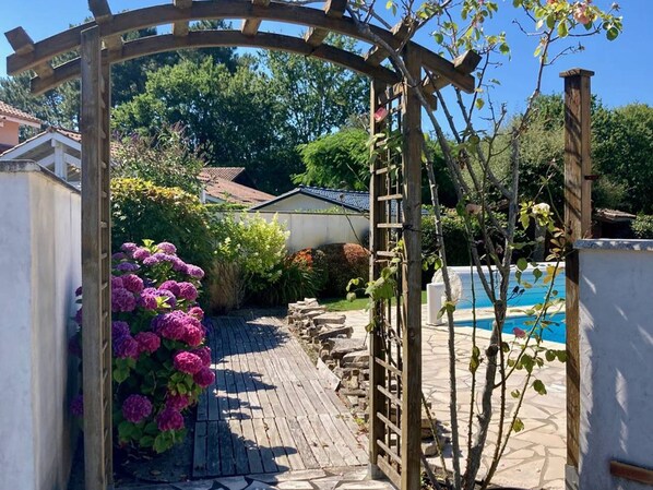 A wooden pergola with a pathway leading to a pool area, surrounded by lush greenery and flowering plants. - House with private pool - 8 guests (Moliets-et-Maa)