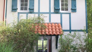 Ett traditionellt hus med korsvirkesfasad, två fönster och fönsterluckor, omgivet av grönska.