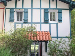 Una casa tradicional con entramado de madera, con dos ventanas y contraventanas, rodeada de vegetación.