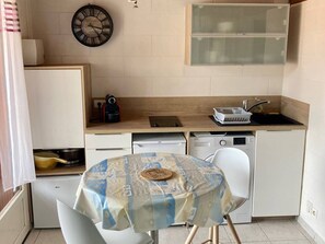 Eine kleine Küche mit einer Uhr, einem Spülbecken, einem kleinen Kühlschrank und einem Tisch mit Tischdecke.