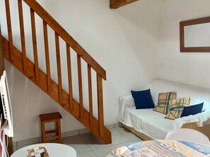 Ein Zimmer mit einer Holztreppe, einem weißen Sofa mit gemusterten Kissen, einem kleinen Holzschemel und einem Tisch mit einem Gesellschaftsspiel.