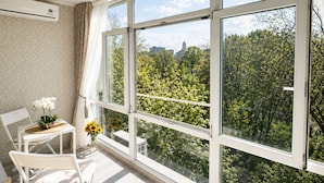 Un balcón con vista a árboles y un edificio, una silla, una mesita con flores y un equipo de aire acondicionado.