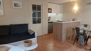 Un espace de vie compact comprenant un canapé, une petite table à manger, un coin cuisine avec un évier et des placards, ainsi qu’un luminaire fixé au mur.