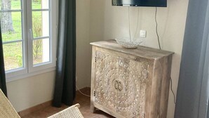 Une pièce avec un téléviseur fixé au mur, un meuble décoratif, un fauteuil en osier et une fenêtre avec des rideaux.