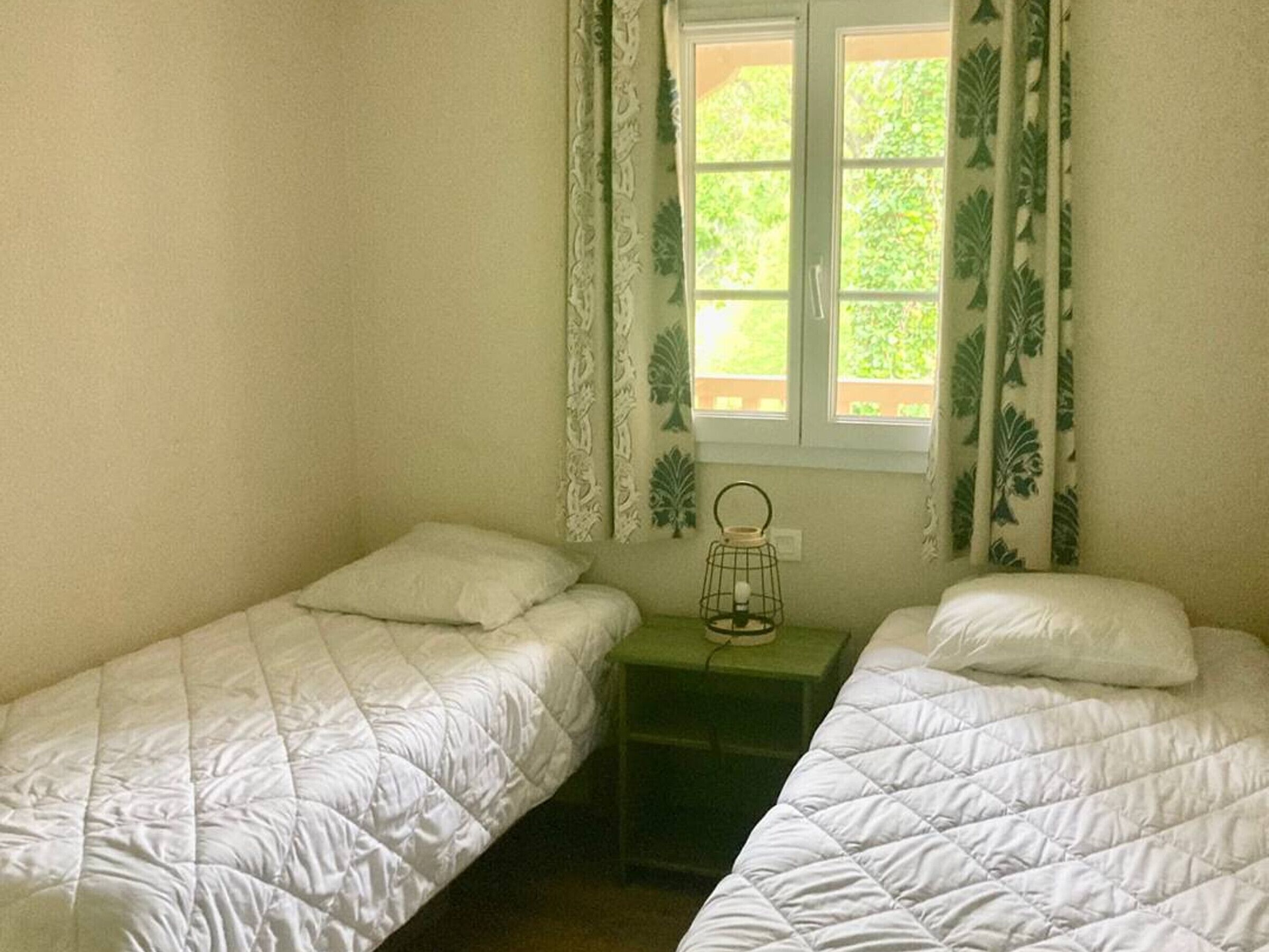 Een kamer met twee eenpersoonsbedden, een kleine tafel met een lamp en een raam met gordijnen.