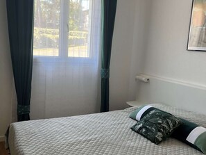 Ein Zimmer mit einem Bett, einem fensterförmigen Ausblick mit Vorhängen, einer Wandleuchte und einem gerahmten Bild.
