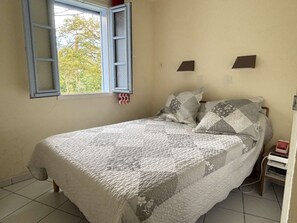 Ein Einzelbett mit Decke, ein Fenster mit geöffnetem Fliegengitter und ein kleiner Nachttisch mit einem Buch.