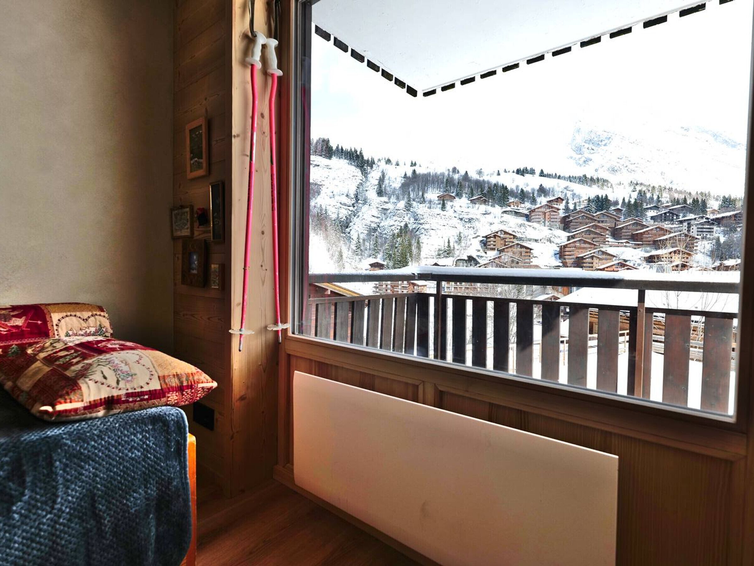 Une pièce avec un plancher en bois, un lit recouvert d’une couverture à motifs, une fenêtre donnant sur des maisons et des montagnes enneigées.