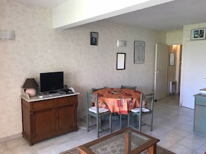Ein Wohnzimmer mit Fernseher, einer Holzeinrichtung, einem Esstisch mit Stühlen und einer Küche im Hintergrund.