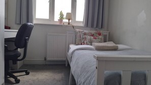 A bedroom with a bed, a desk with a chair, a radiator, a window with curtains, and a small potted plant. - Charming 4-bedroom Apartment in the Heart of London (London)