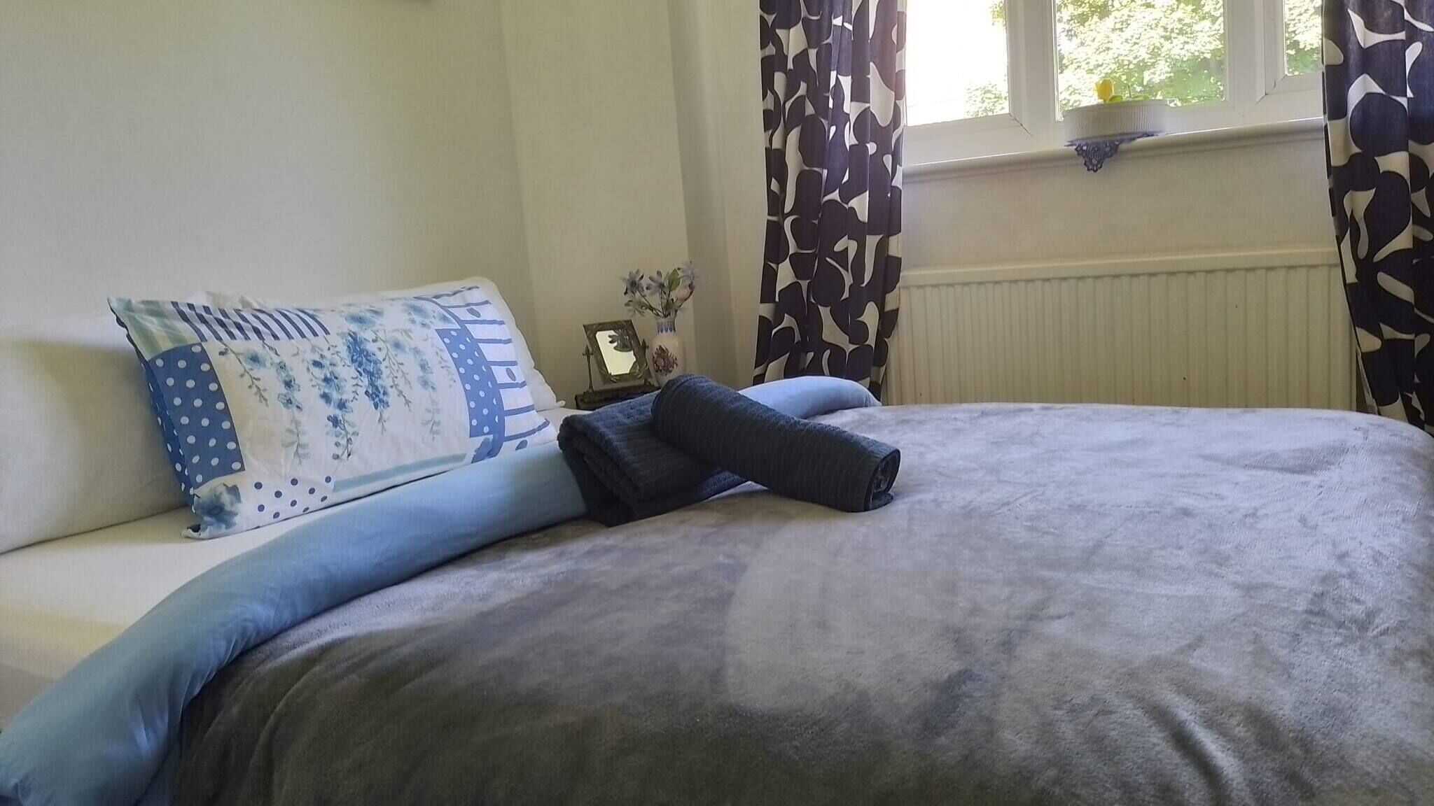 A neatly made bed with a blue and white patterned pillow, a rolled-up towel, and a grey blanket.