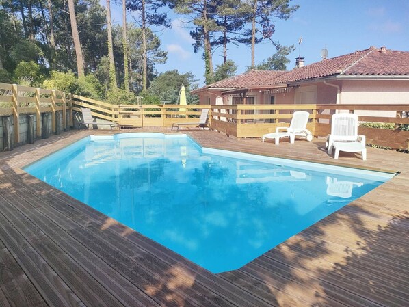 Una piscina rettangolare con un pontile in legno, circondata da una recinzione di legno e sedie bianche.