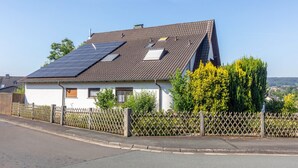 Een huis met een schuin dak, zonnepanelen, een omheinde tuin en een heldere lucht.