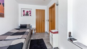 Een slaapkamer met een bed, een klein tafeltje, een stoel, een houten deur en een wandplank met een apparaat.