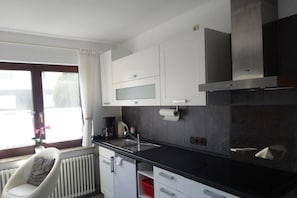 Une cuisine moderne avec des meubles blancs, un plan de travail noir, une cuisiniĂšre encastrĂ©e et une fenĂȘtre avec des rideaux.