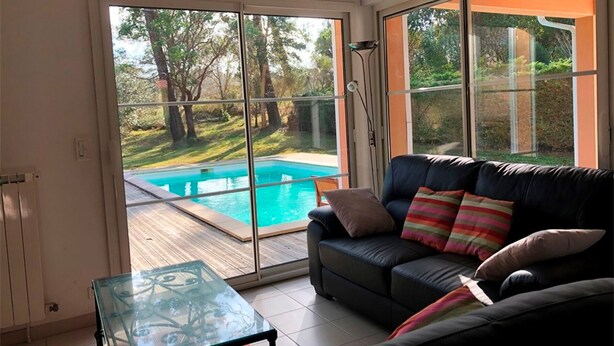 Un salon avec un canapé en cuir noir, une table basse en verre et une vue sur une piscine, visible à travers de grandes portes vitrées.