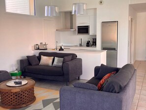 Ein modernes Wohnzimmer mit grauem Sofa, einem kleinen Couchtisch und einer Küchenzeile mit weißen Schränken und Edelstahlgeräten.